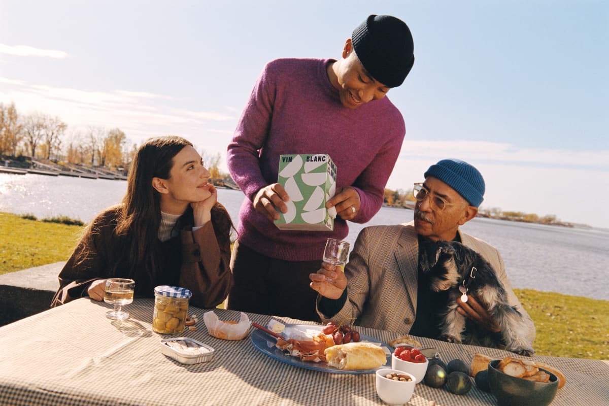 Three people outside, sitting at a bench in a park drinking Ami Ami boxed wine.